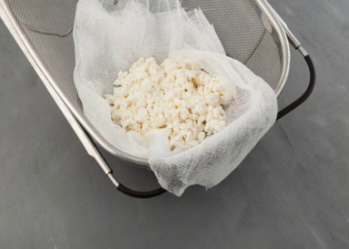 Застелите дуршлаг марлей и переложите на нее всплывшие сырные зерна. Сыр накройте свободными концами ткани и оставьте до остывания.